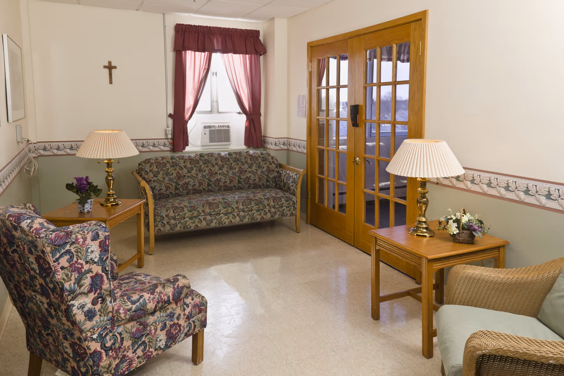 A small sitting area with floral patterned couch and armchair, a wicker chair with a cushion, two wooden side tables each with a brass lamp, a window with maroon curtains and an air conditioning unit, and wooden French doors. A cross hangs on the wall above the couch.