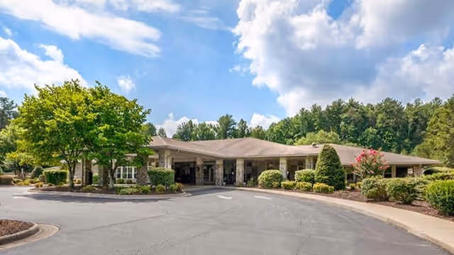 Exterior view of a single-story senior living facility building with a covered entrance, surrounded by well-maintained landscaping including trees, bushes, and flowering plants under a partly cloudy sky.