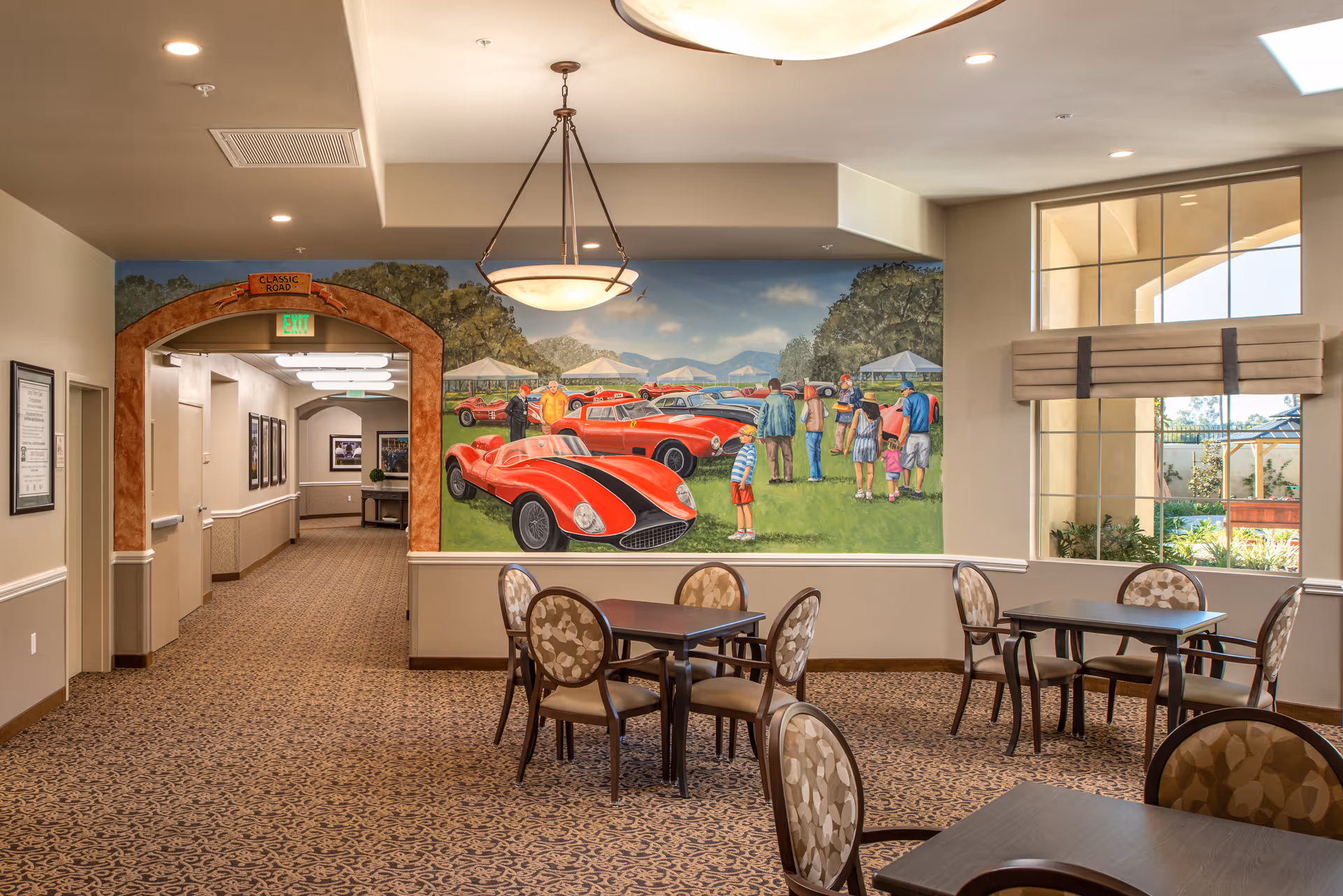 Interior view of a common area in a senior living facility with several tables and chairs. A large mural on the wall depicts a classic car show with people admiring red vintage cars on a grassy field. There is a hallway with framed pictures and an exit sign above an archway labeled 'Classic Road'. A large window lets in natural light, showing some outdoor greenery.