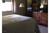 A bedroom with a large bed covered in a gray bedspread, a window with curtains letting in natural light, a floor lamp beside the bed, and a wooden dresser with a flat-screen TV on top. A mirror above the dresser reflects part of the room.