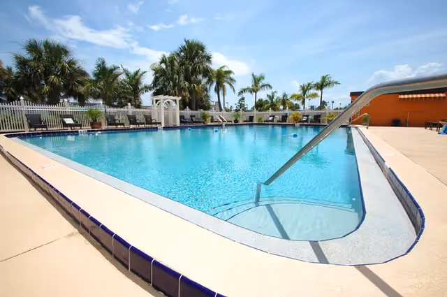 Outdoor swimming pool with clear blue water, surrounded by a concrete deck and lounge chairs. Palm trees and a white fence are visible in the background under a partly cloudy sky.