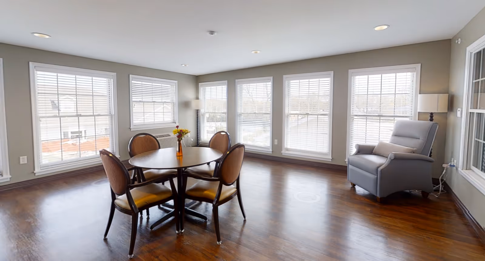A bright room with large windows featuring white blinds, a round wooden table with four cushioned chairs, a small vase with flowers on the table, a gray armchair with a pillow, and two floor lamps in the corners. The room has wooden flooring and neutral-colored walls.
