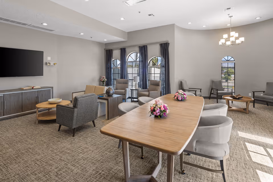 A spacious senior living community room with a long wooden table decorated with flower arrangements, surrounded by cushioned chairs. The room features multiple seating areas with armchairs and small tables, large windows with arched tops and dark curtains, a wall-mounted flat-screen TV, and neutral-colored walls and carpet. A modern chandelier hangs from the ceiling, providing warm lighting.