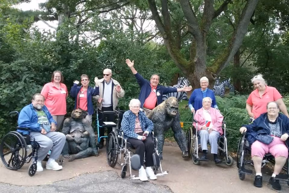 A group of elderly people, some in wheelchairs, and caregivers posing outdoors in front of greenery and tree sculptures of a gorilla and a chimpanzee. The group is smiling and waving, enjoying a day outside.
