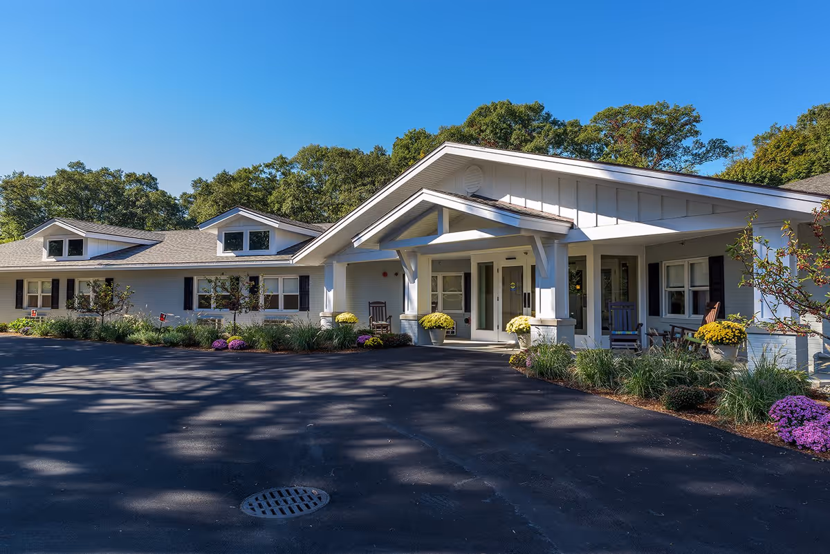 Exterior view of a single-story senior living facility building with a covered entrance, white siding, and a paved driveway. There are potted plants with yellow and purple flowers near the entrance and small trees and shrubs along the front of the building. The sky is clear and blue, and there are trees in the background.
