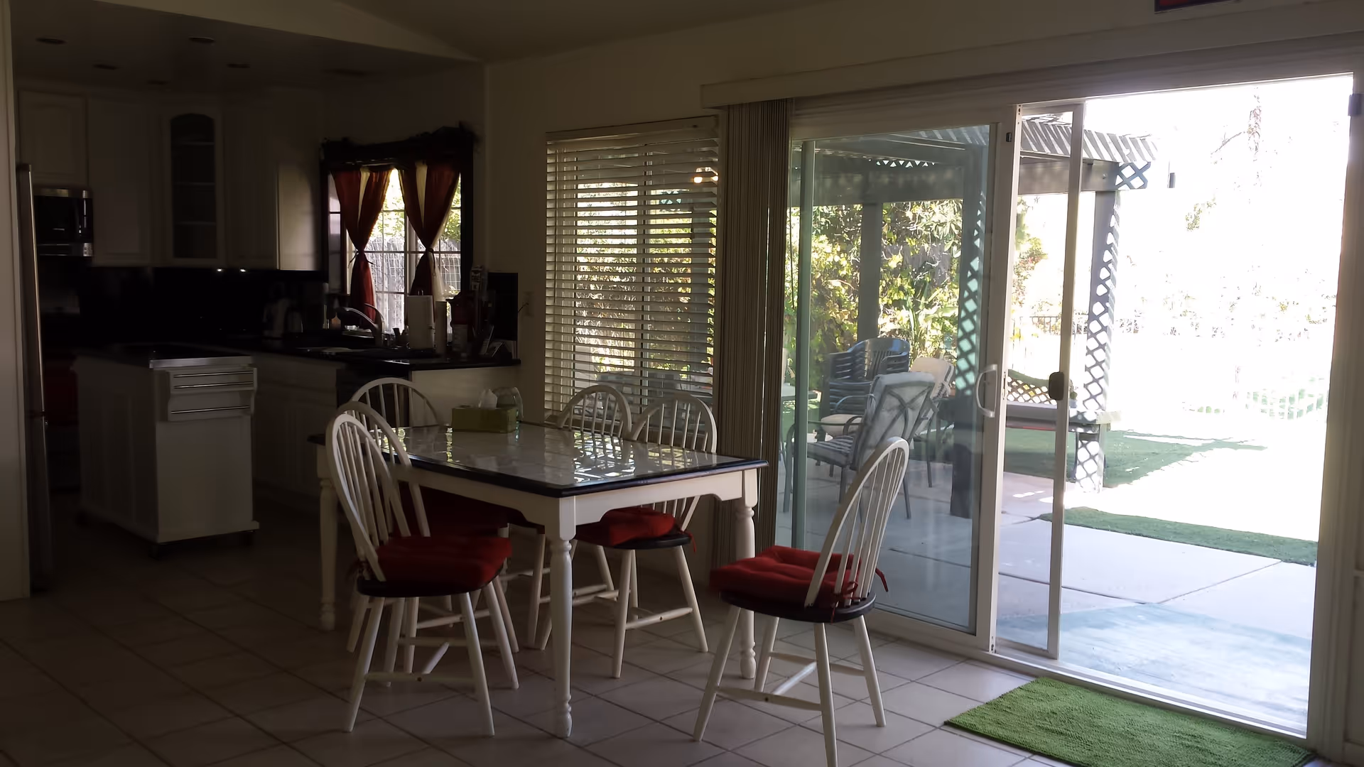 Interior view of a dining area with a white table and six white chairs with red cushions. The dining area is adjacent to a kitchen with white cabinets and a small island. There is a sliding glass door leading to an outdoor patio with chairs and greenery visible outside.