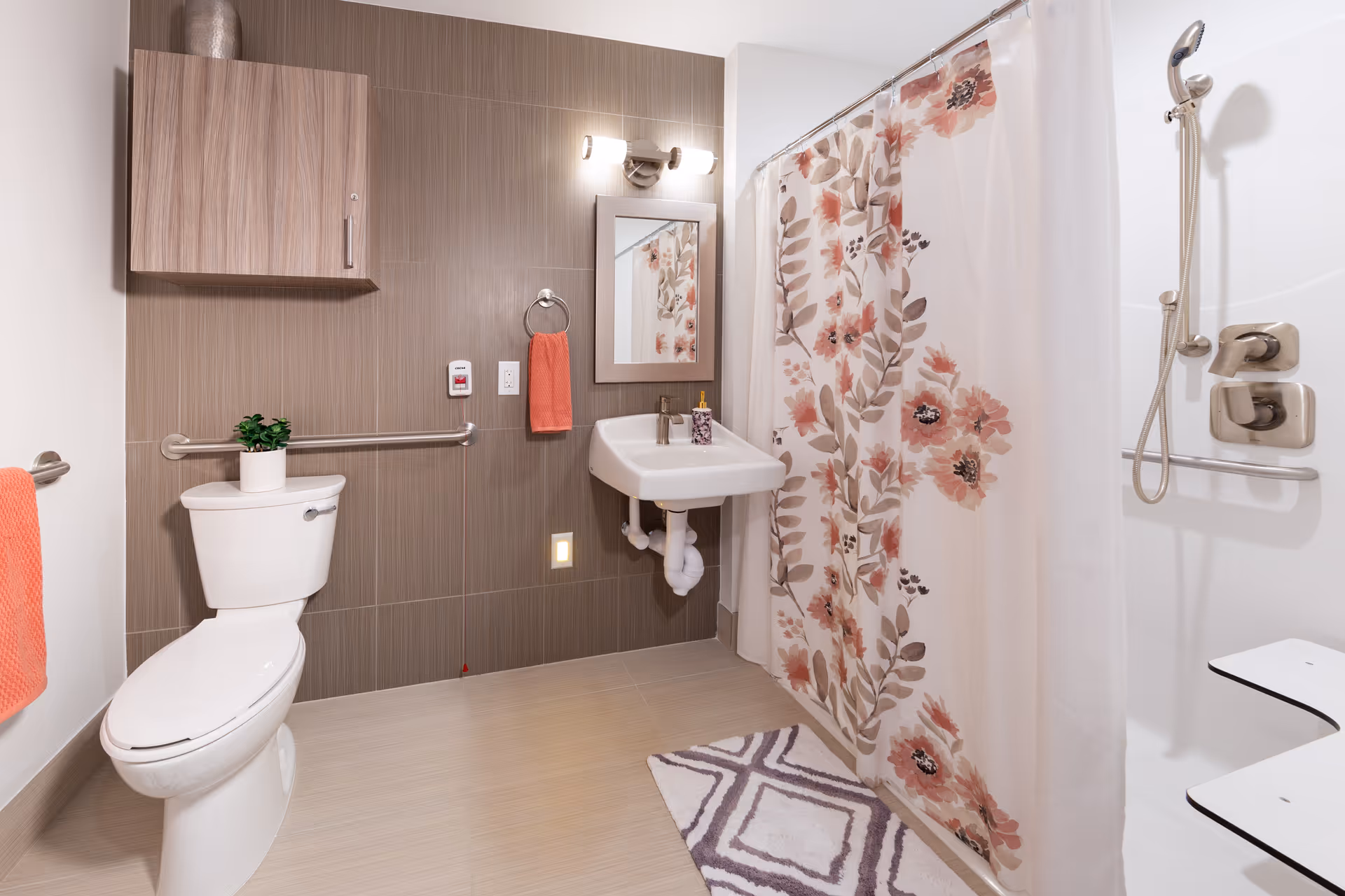 A clean and modern bathroom with a white toilet, a small potted plant on the toilet tank, a wall-mounted sink with a soap dispenser, a mirror above the sink, and a shower area with a floral shower curtain and a handheld showerhead. The bathroom features beige tiled walls and floor, a wooden cabinet mounted on the wall, and orange towels hanging on towel rings.