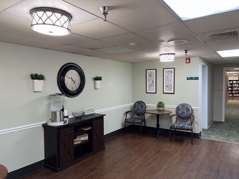 A hallway corner in a senior living facility with light green walls and dark wood flooring. There is a dark wood cabinet with a water dispenser, a large round wall clock, and two small potted plants mounted on the wall. To the right, there is a small round table with two patterned chairs and a small plant on the table. Two framed artworks hang on the wall above the table. The hallway extends to the right, leading to a room with bookshelves visible in the distance.