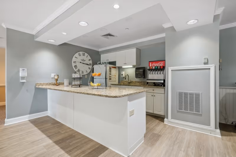 Interior view of a senior living facility kitchen area with a granite countertop island, white cabinets, a large wall clock, a Coca-Cola soda dispenser, and a tiered fruit stand with oranges and bananas.