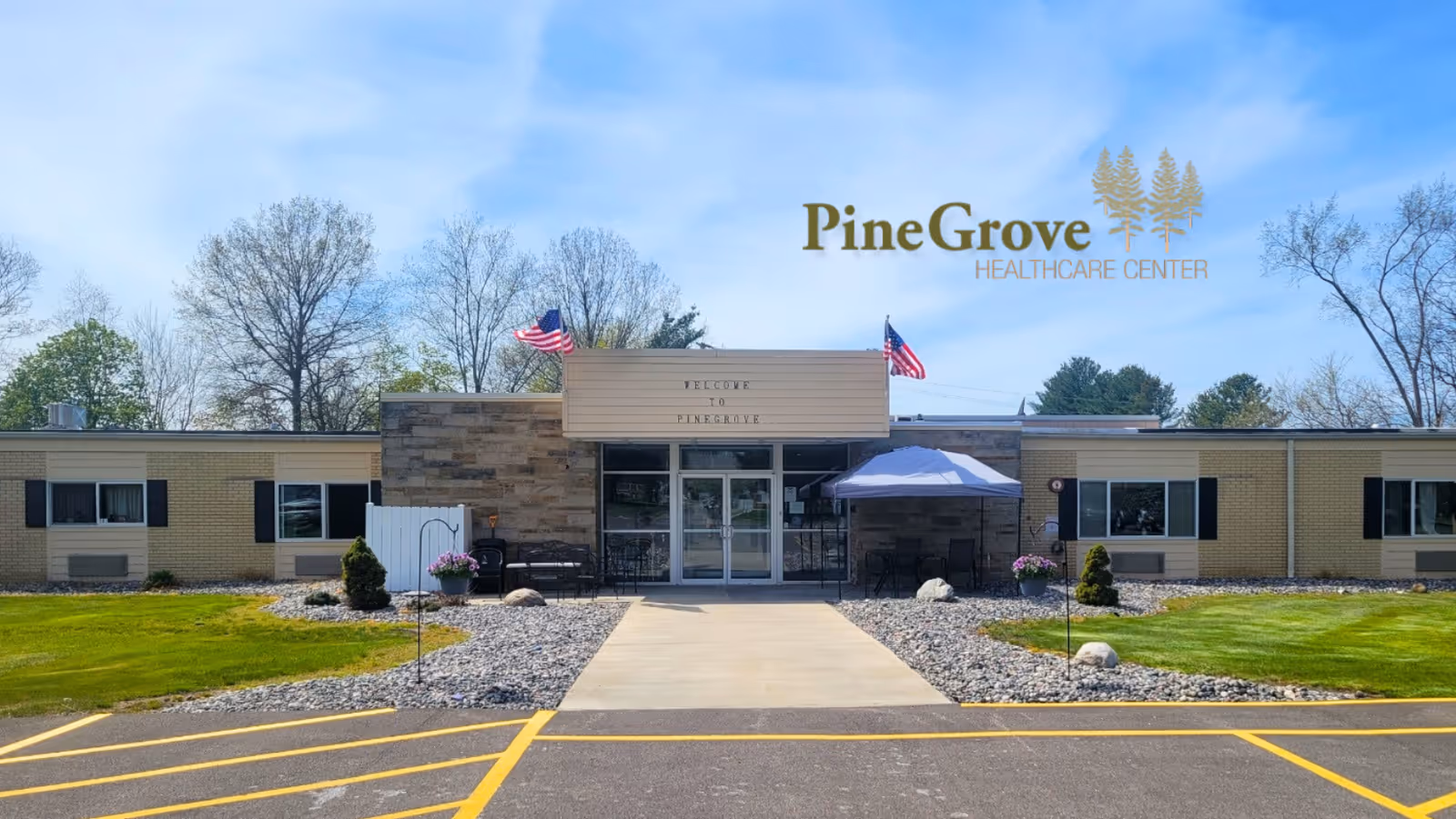 Front exterior view of Pine Grove Healthcare Center building with a pathway leading to the entrance, two American flags, some outdoor seating with an umbrella, and trees in the background under a blue sky.