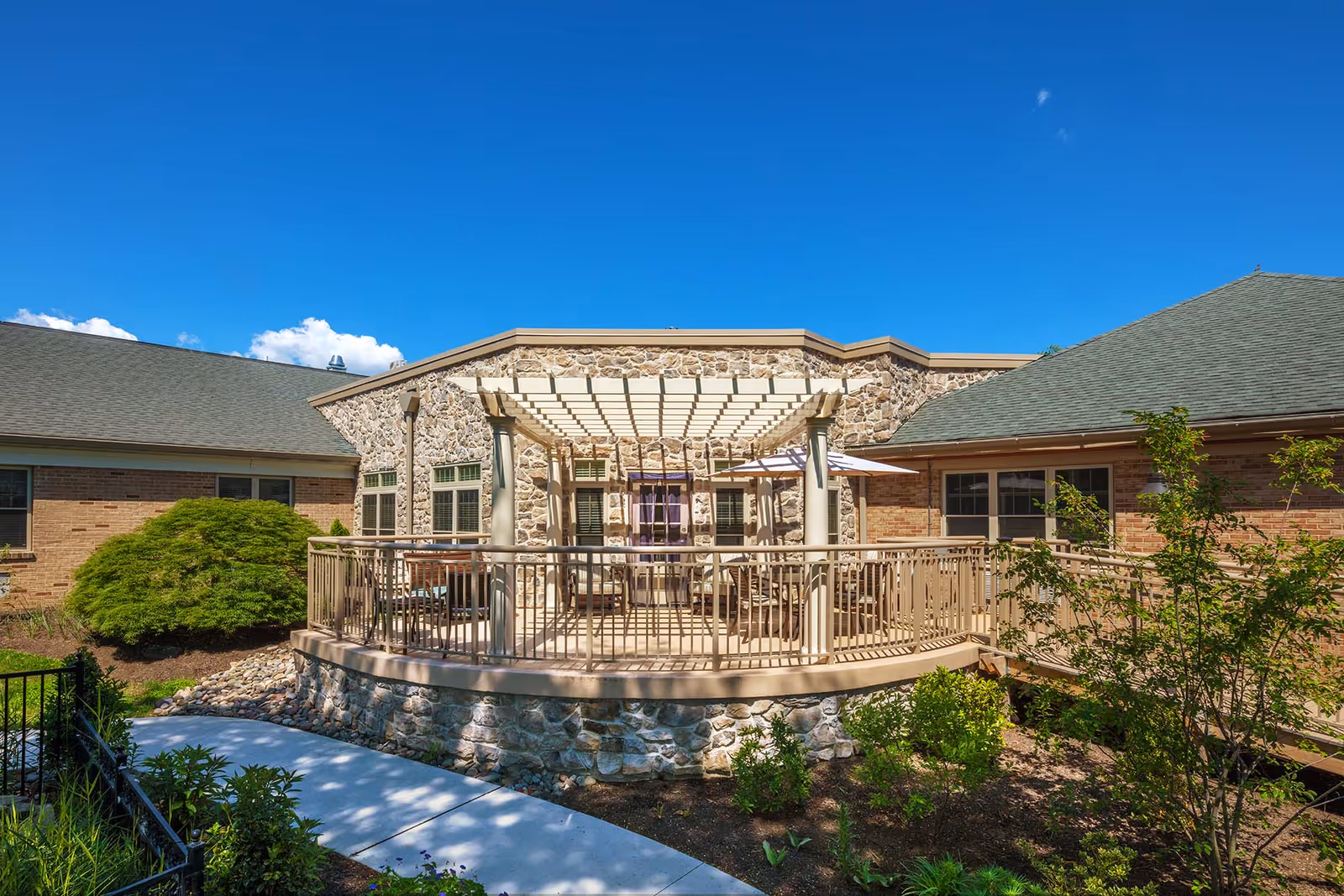 Outdoor patio area at Artman Lutheran Home featuring a stone and brick building with a pergola-covered seating area, surrounded by greenery and a clear blue sky.