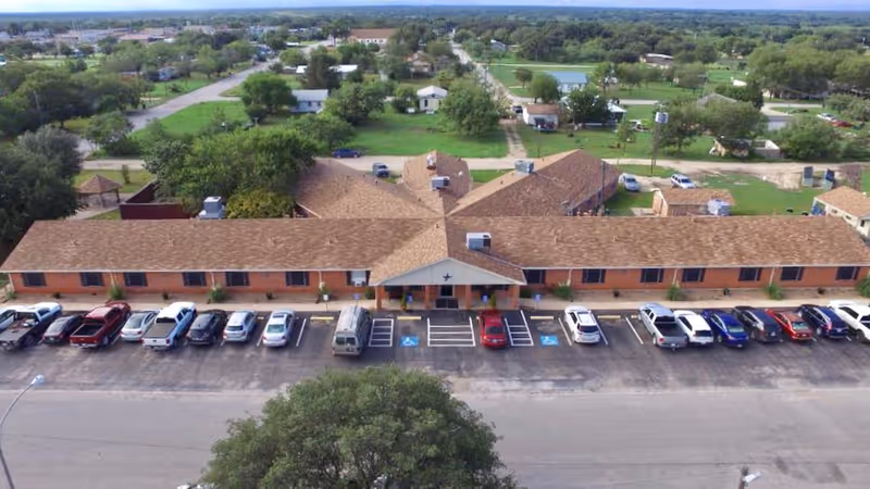 Aerial view of Concho Health and Rehabilitation Center showing a single-story building with a brown shingle roof and multiple parked cars in front. The building is surrounded by green lawns, trees, and a residential neighborhood in the background under a clear sky.