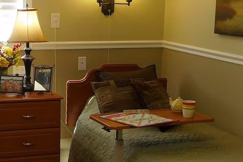 A cozy bedroom in a senior living facility featuring a single bed with multiple pillows and a green quilt. A wooden bedside table holds a lamp, framed photos, and a vase with flowers. A small adjustable table on the bed has a Scrabble game in progress and a cup. The walls are painted beige with white trim, and a painting is partially visible on the right wall.