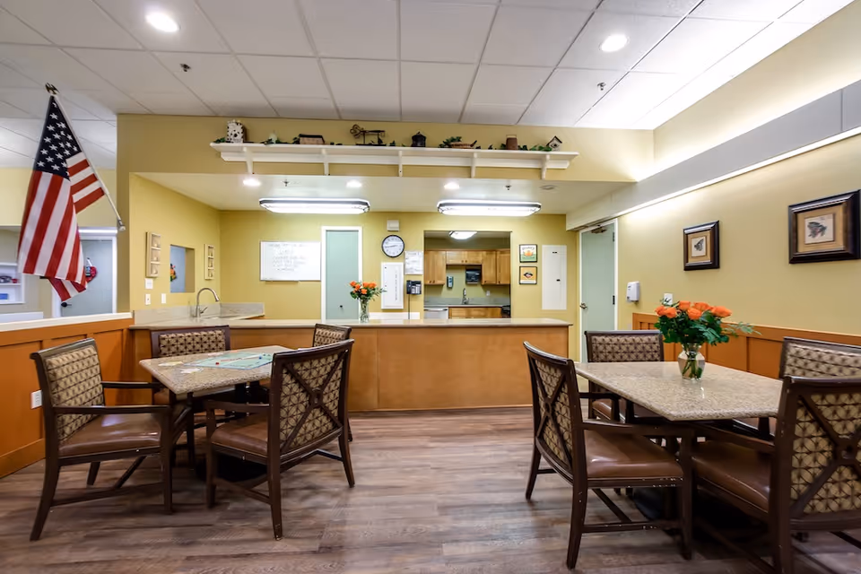 Interior view of a senior living facility dining area with two tables, each surrounded by four cushioned chairs. The room has wooden flooring, yellow walls with wood paneling, and a kitchen counter with a sink and cabinets in the background. An American flag is displayed on the left side, and there are framed pictures and a vase with orange flowers on the tables.