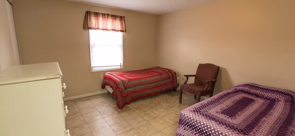 A simple bedroom with two single beds covered with crocheted blankets, one red and one purple. There is a wooden chair between the beds and a white dresser on the left side. A window with a plaid valance lets in natural light, and the floor is tiled.