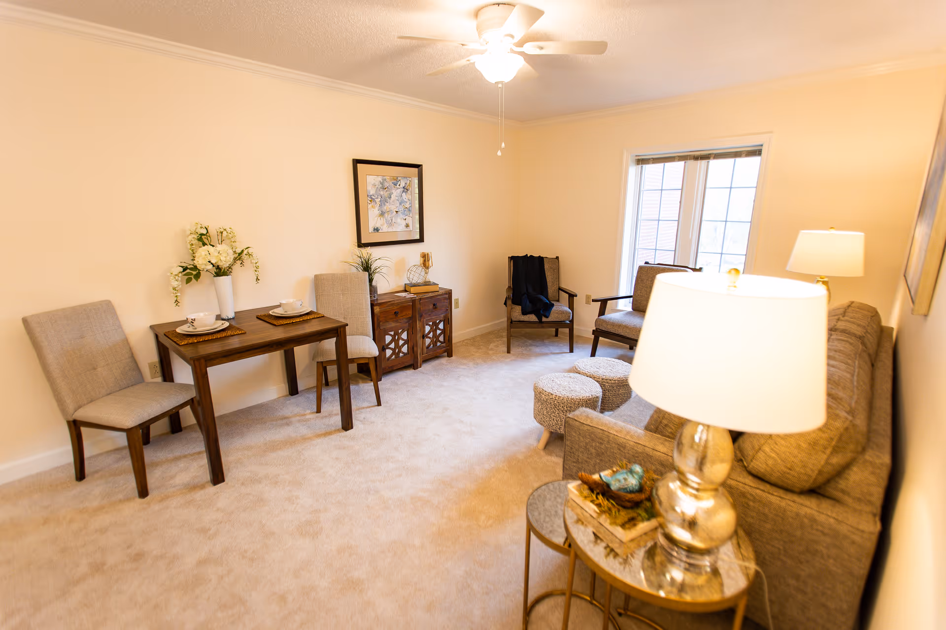 A cozy living room with beige walls and carpet. The room features a small wooden dining table with two chairs, a sideboard with decorative items, two armchairs near a window, a gray sofa, two round ottomans, and a side table with a lamp and decorative items. A ceiling fan with a light is mounted on the ceiling.