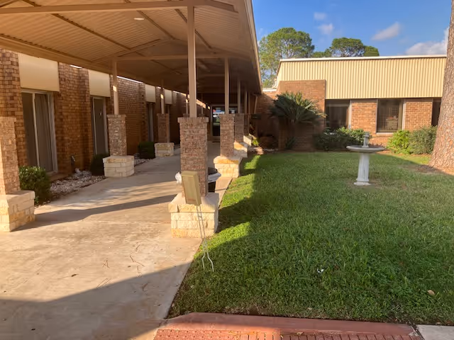 Covered brick walkway alongside a grassy courtyard with a birdbath in front of a low brick building.