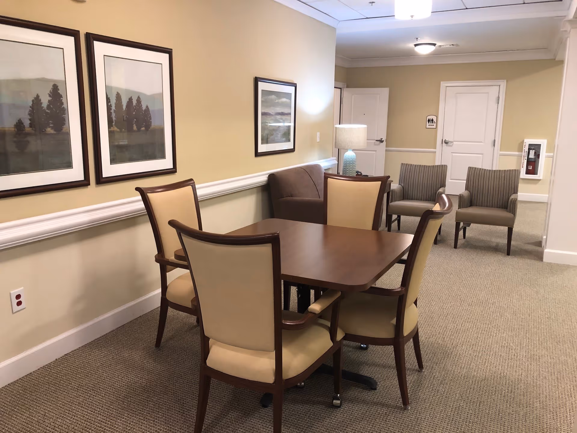 A cozy interior room with a wooden table surrounded by four beige cushioned chairs. In the background, there is a brown loveseat, two striped armchairs, a table lamp with a blue base, and framed landscape artwork on the beige walls. The room has carpeted flooring and two white doors visible in the back.