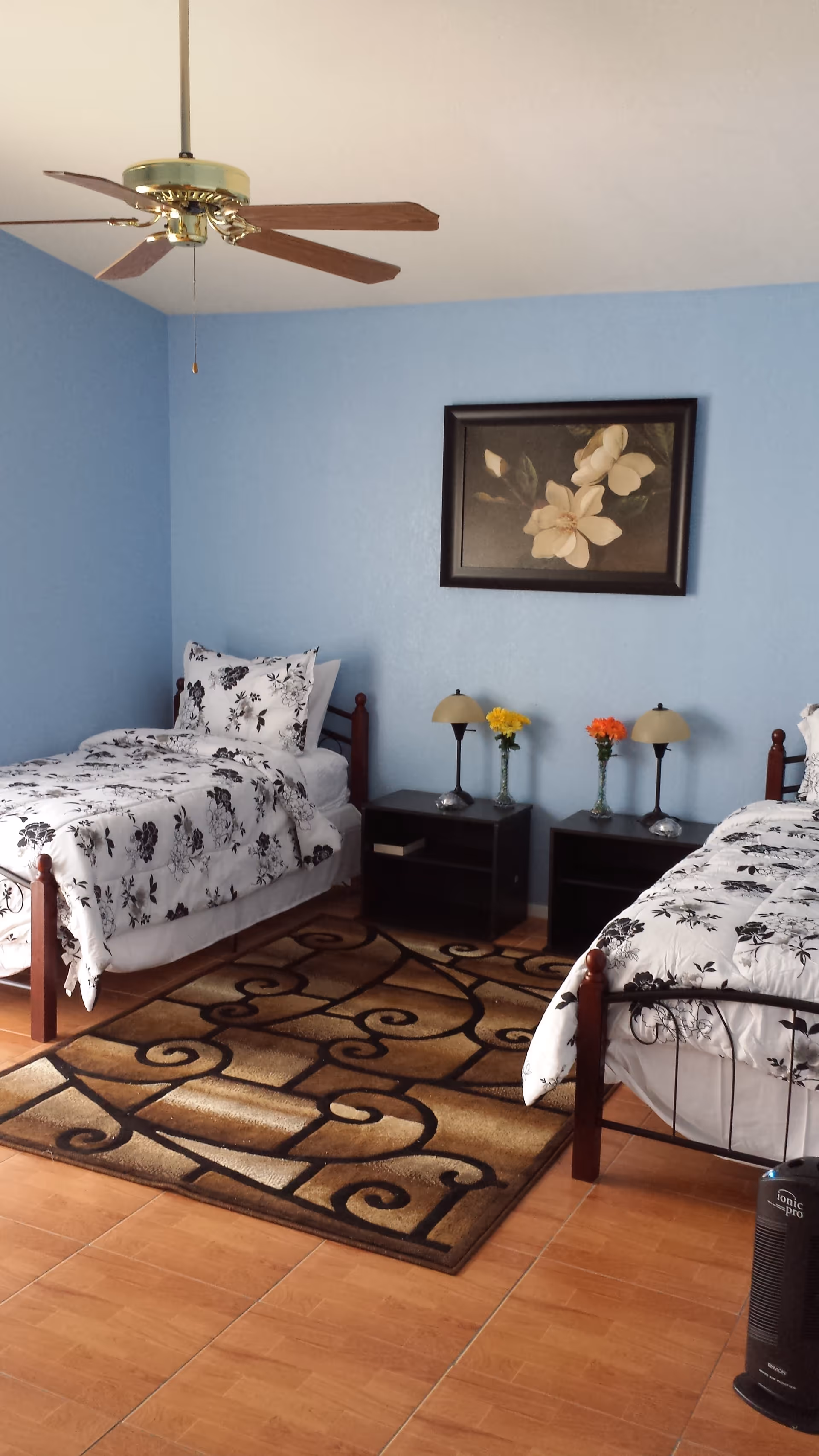 A bedroom with two single beds featuring black and white floral bedding. Between the beds are two black nightstands, each with a small lamp and a vase of flowers. A framed floral painting hangs on the light blue wall above the nightstands. The floor is tiled with a brown patterned area rug in the center. A ceiling fan with wooden blades is visible above.