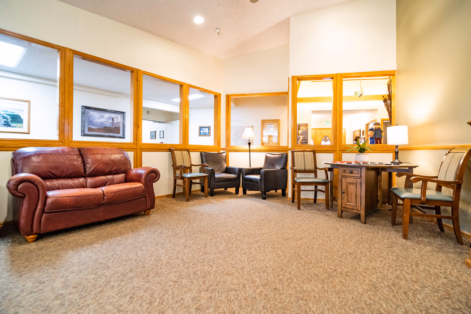A cozy seating area in a memory care facility with a red leather sofa, two black leather chairs, and several wooden chairs arranged around a small wooden table with a lamp and decorative items. The walls have large windows with wooden frames, and framed pictures hang on the walls.