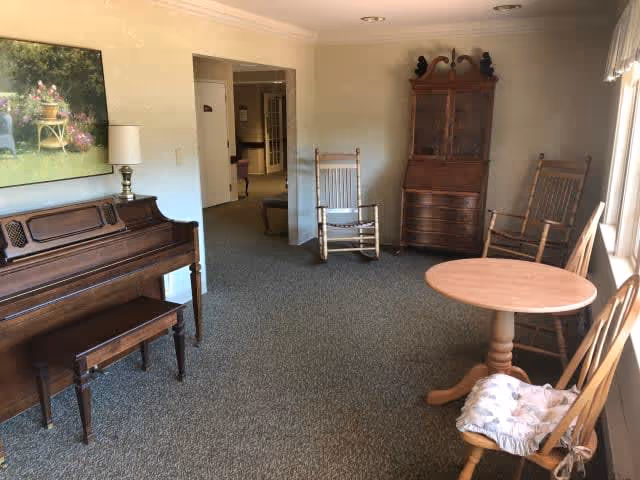 A cozy living room area with a wooden piano and bench on the left, two wooden rocking chairs, a round wooden table, and a wooden cabinet against the far wall. The room has carpeted flooring and a large window on the right letting in natural light.
