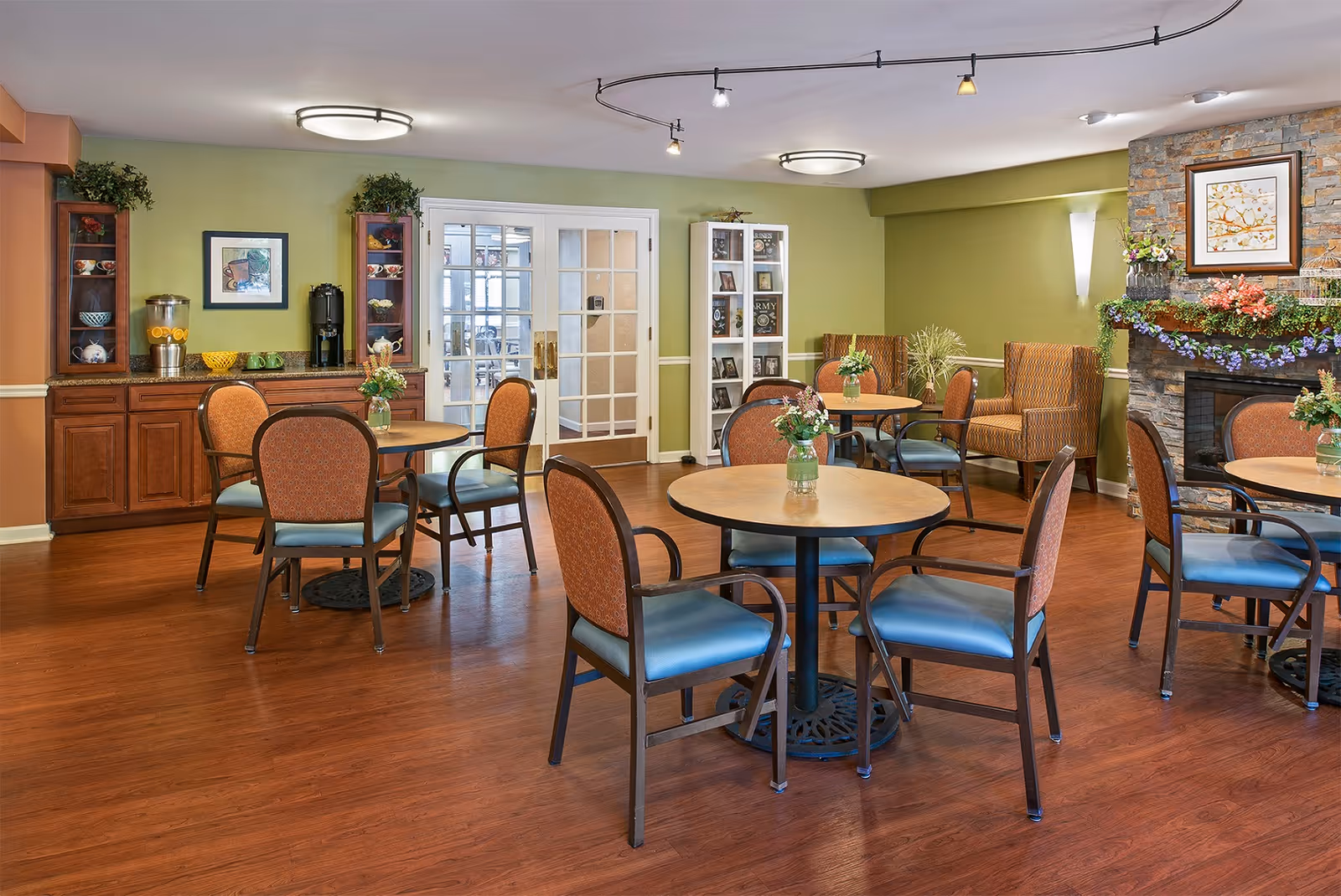 A cozy dining area in a senior living facility with round tables and chairs arranged on a wooden floor. The walls are painted green and orange, decorated with plants, framed artwork, and a stone fireplace adorned with flowers. There are glass-front cabinets and a beverage dispenser on a wooden counter against one wall.