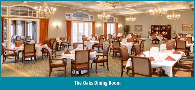 A spacious dining room with multiple round and rectangular tables covered with white tablecloths and set with red napkins. The room features large windows with red curtains, chandeliers hanging from a coffered ceiling, and a carpeted floor. There are wooden chairs around the tables and a wooden cabinet against the far wall.
