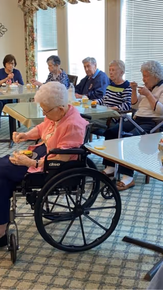 Several people seated around dining tables in a communal room, with one woman in a wheelchair eating a snack.