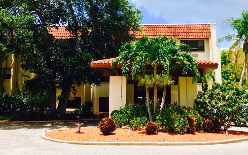 Front exterior view of a building with a red tiled roof, surrounded by lush green trees and shrubs, with a circular driveway and a small landscaped island featuring palm trees and bushes.