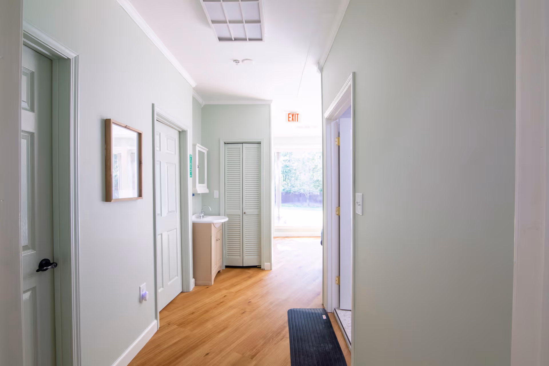 A bright hallway with light green walls and wooden flooring. There are several doors along the hallway, a small sink with a mirror above it, and a closet with louvered doors at the end. An exit sign is visible above a door leading outside, where greenery can be seen through a window.