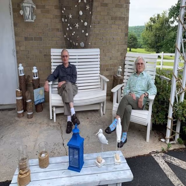 Two elderly individuals sitting on white wooden chairs on a porch with a brick wall behind them. There are decorative items including a blue lantern, glass candle holders, and bird figurines on a white wooden table in front of them. The porch overlooks a green outdoor area with trees and a white fence.