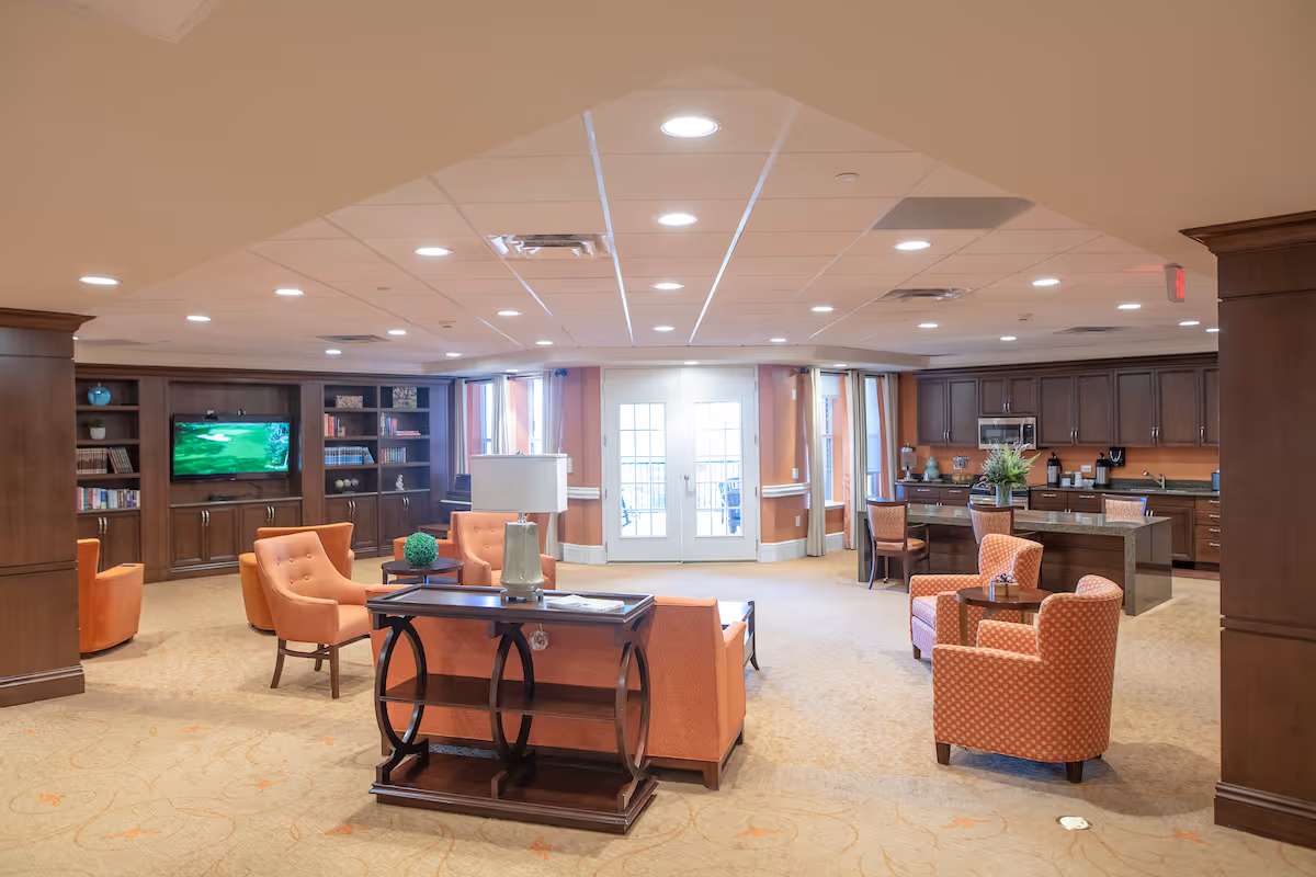 A spacious senior living common area with orange upholstered chairs and sofas arranged around small tables. There is a wooden console table with a lamp in the foreground. The back wall features built-in wooden shelves filled with books and a flat-screen TV. To the right, there is a kitchen area with dark wood cabinets, a microwave, and a granite countertop island with chairs. Large windows and glass doors let in natural light.