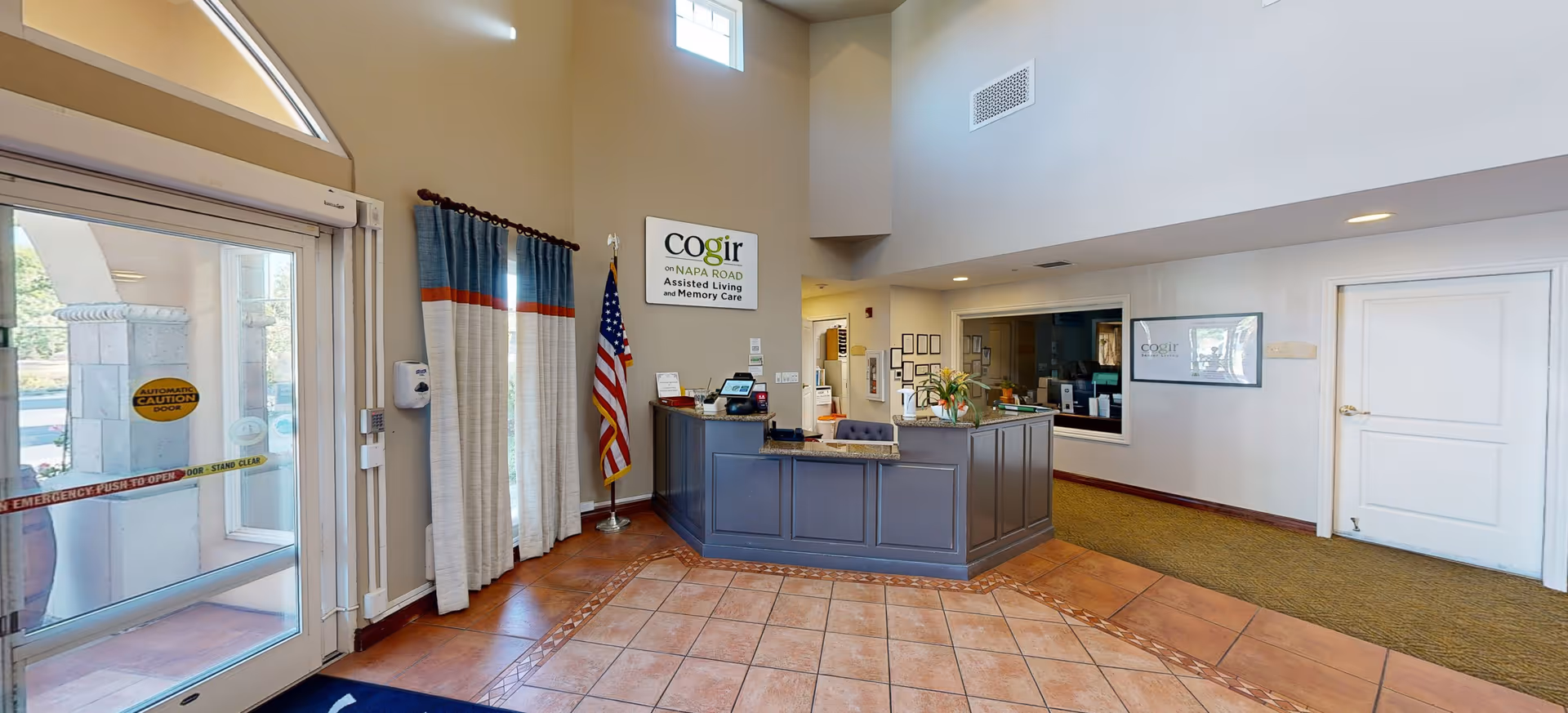 Reception area of Cogir of Sonoma Plaza assisted living facility with a front desk, an American flag, a sign on the wall, tiled floor, and a glass entrance door with caution tape.