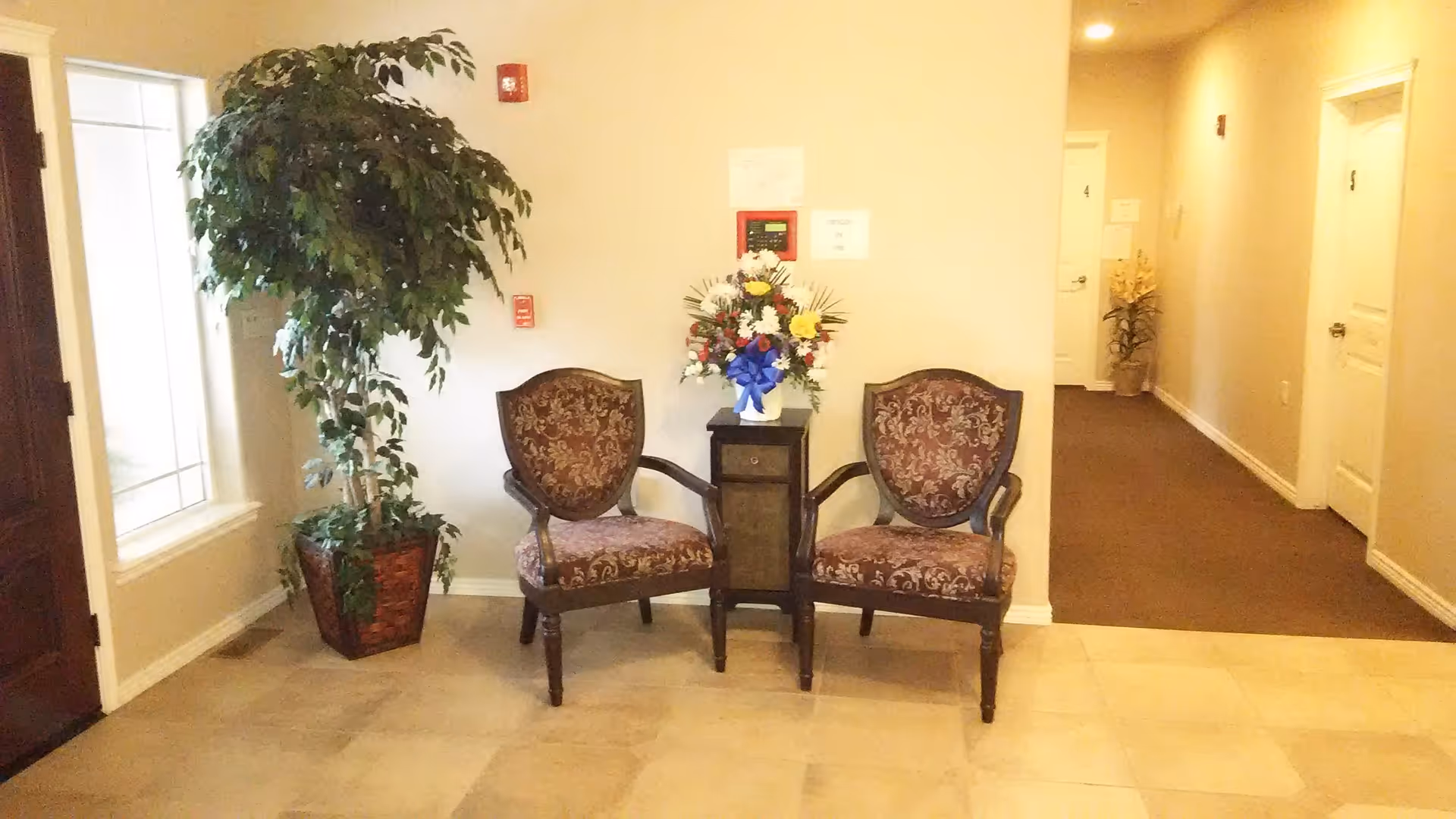A small seating area in a hallway with two ornate upholstered chairs and a small table between them holding a floral arrangement with a blue ribbon. To the left is a large potted plant near a window, and to the right is a hallway with doors numbered 4 and 5. The floor is tiled near the seating area and carpeted in the hallway.