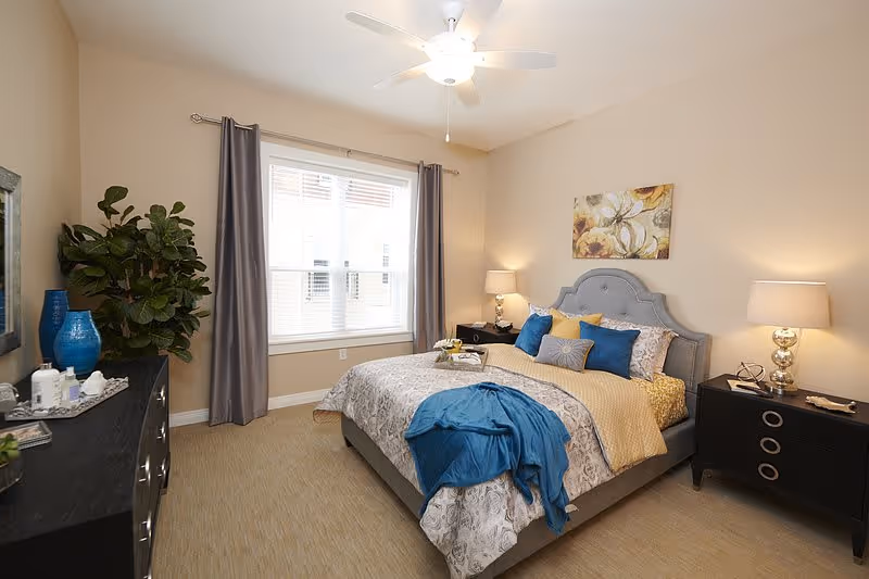 A well-lit bedroom with a large bed featuring a gray upholstered headboard, decorated with multiple pillows in blue, yellow, and gray tones. A blue throw blanket is draped over the bed. On either side of the bed are black nightstands with lamps. A window with gray curtains lets in natural light. A dresser with decorative items and a large green plant are visible on the left side of the room. A ceiling fan with a light is mounted on the ceiling.