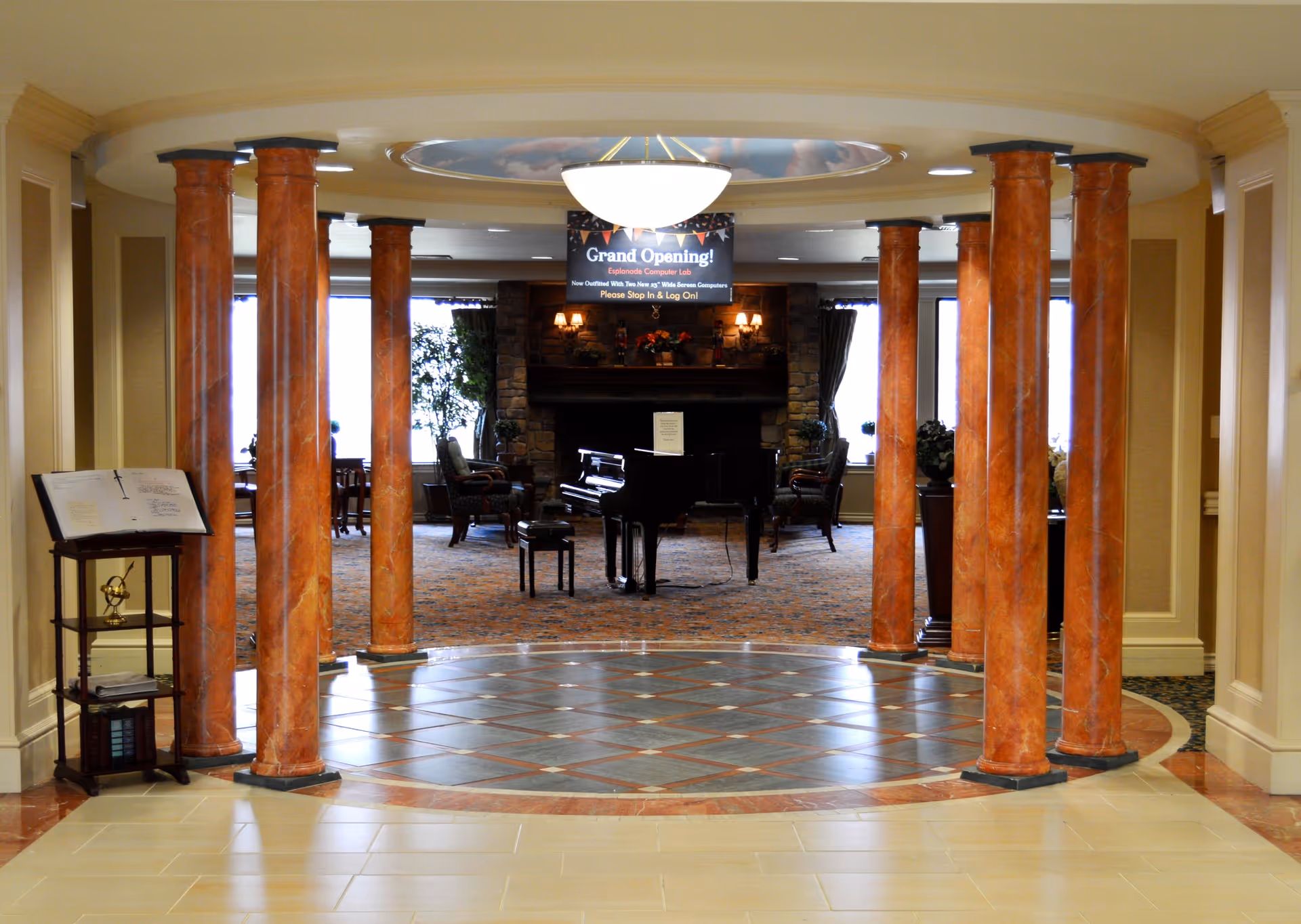 Interior view of a senior living facility featuring a circular area with tall orange marble columns surrounding a tiled floor. Beyond the columns is a room with a grand piano, chairs, and a stone fireplace. A sign hanging from the ceiling announces the grand opening of the Esplanade Computer Lab.