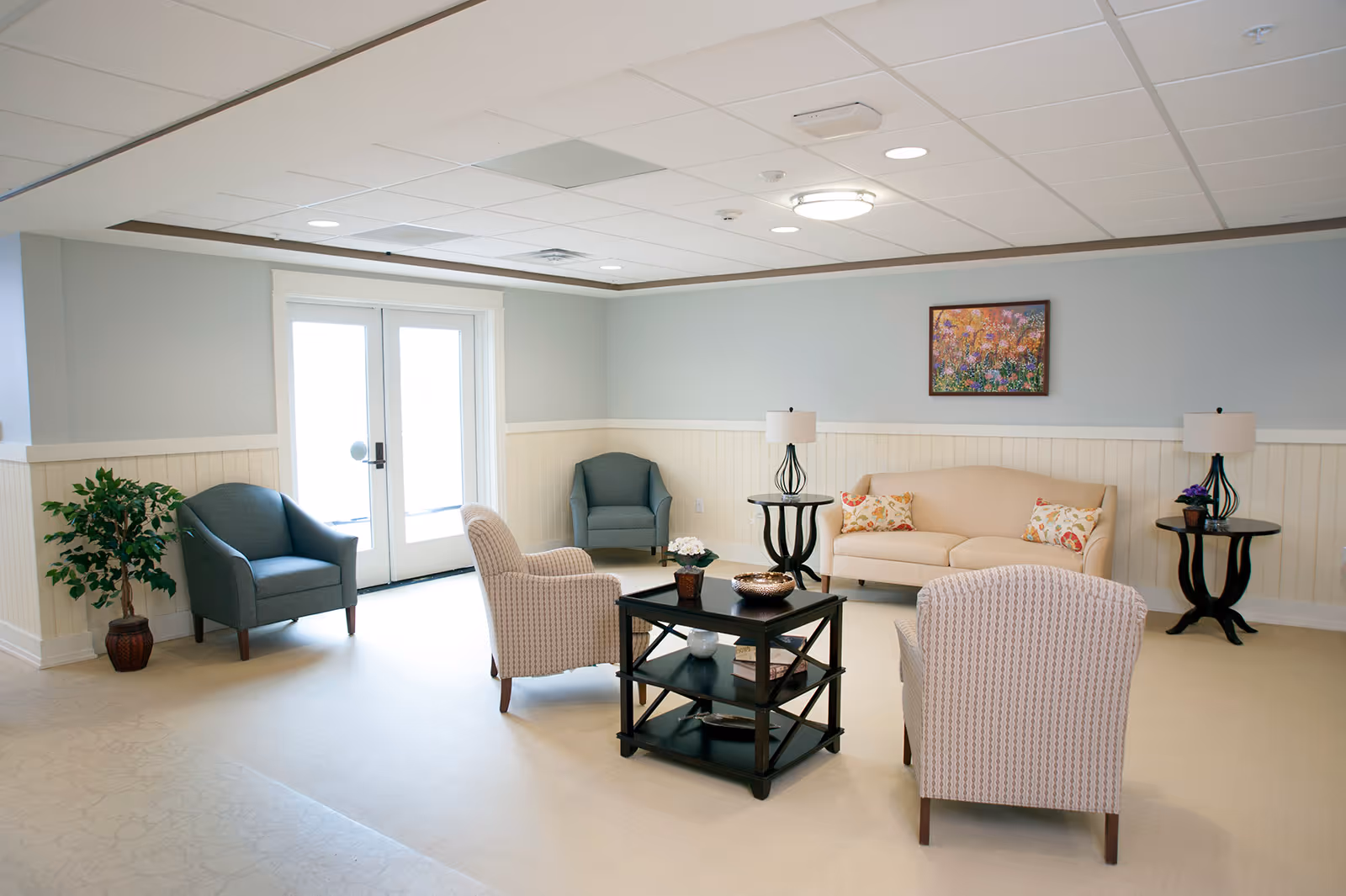 Bright lounge with a beige sofa, several armchairs around a central coffee table, side tables and double glass doors.