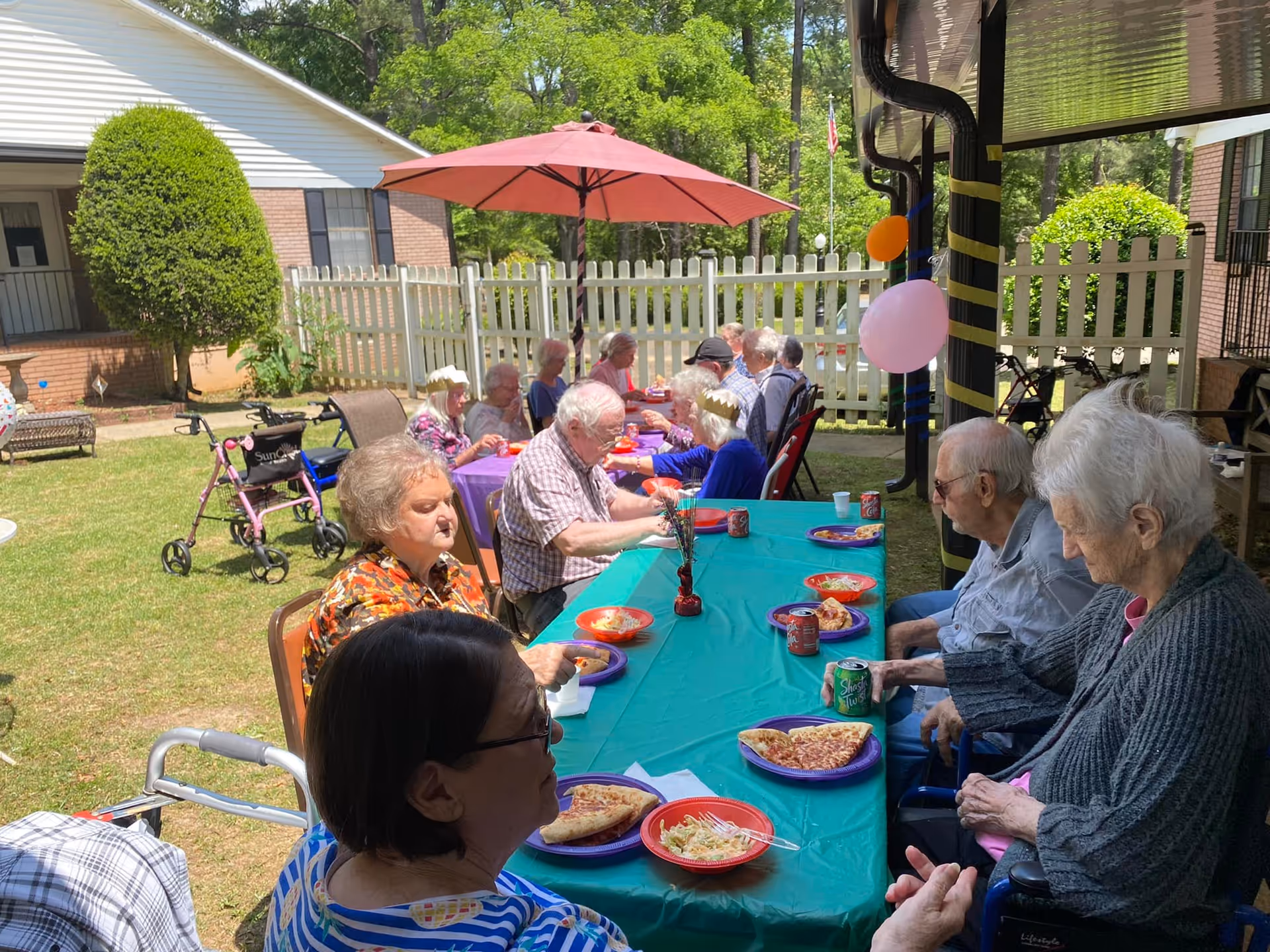 A group of elderly people sitting outdoors around a long table covered with a green tablecloth, eating pizza and salad. The table is set under a red umbrella and decorated with balloons. The setting is a fenced yard with grass and trees, adjacent to a brick building.