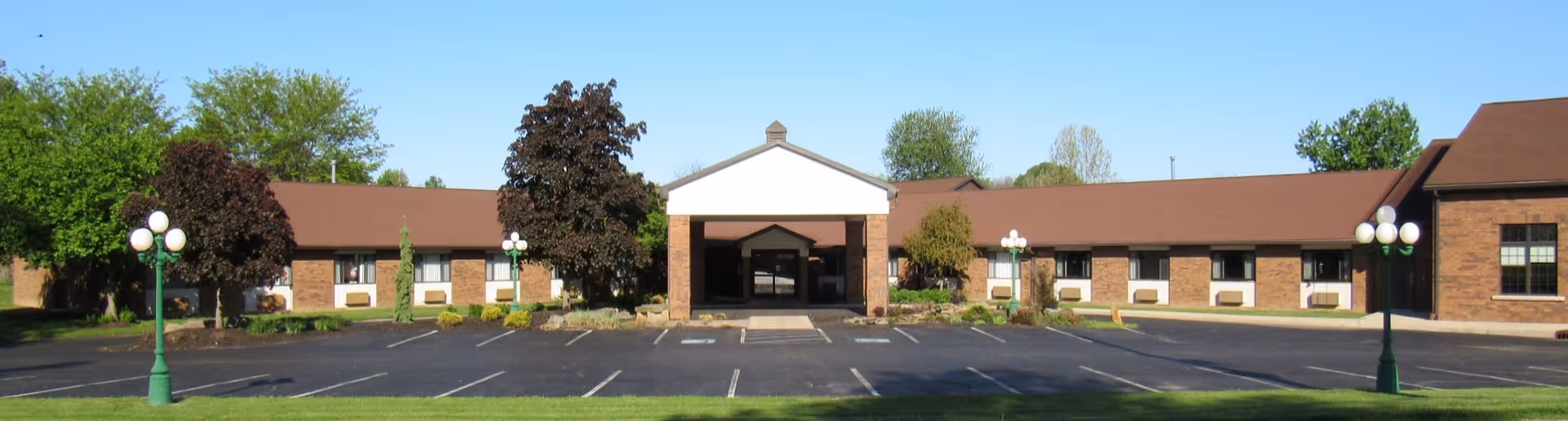 Front exterior view of Century Villa Health Care & Rehabilitation building with a brown roof, brick walls, multiple windows, and a central entrance under a white triangular pediment. The parking lot in front is empty, and there are green lamp posts and trees around the building.