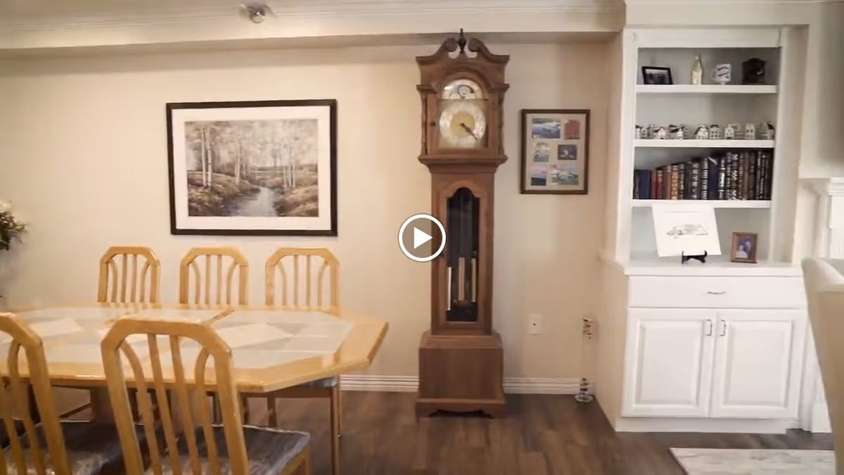 A dining area featuring a wooden dining table with six matching chairs. On the wall behind the table, there is a framed landscape painting and a tall grandfather clock. To the right, there is a white built-in bookshelf with books, small decorative items, and framed pictures. The floor is wooden, and the room has a cozy, traditional style.