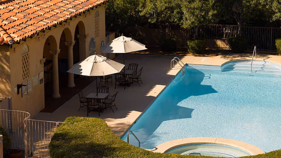 Outdoor swimming pool area with clear blue water, surrounded by a patio with tables, chairs, and umbrellas. The building adjacent to the pool has a tiled roof and arched columns. There are bushes and trees in the background providing shade.