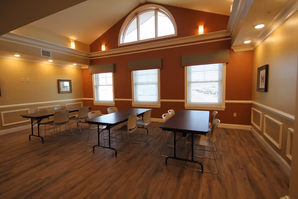 A well-lit dining room with three rectangular tables, each surrounded by white chairs. The room features wood flooring, beige and rust-colored walls with white trim, three large windows with blinds, and a large arched window near the ceiling. Wall sconces provide additional lighting.