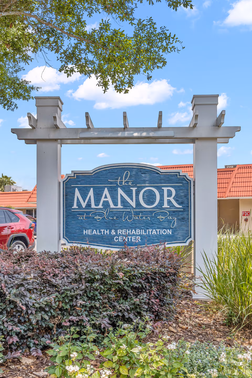 Outdoor view of a large white wooden sign for The Manor at Blue Water Bay Health & Rehabilitation Center, surrounded by bushes and plants with a red-roofed building and blue sky with clouds in the background.