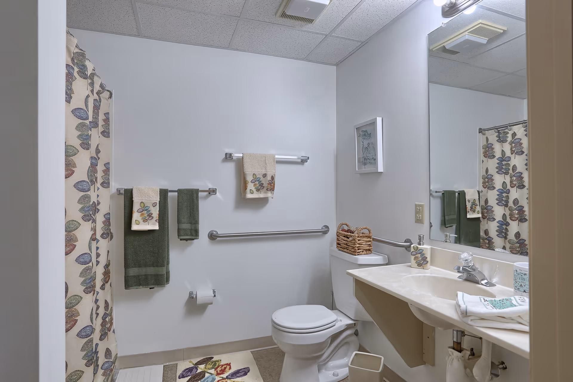 Accessible bathroom with a toilet, sink and mirror, grab bars, floral shower curtain, and green towels.