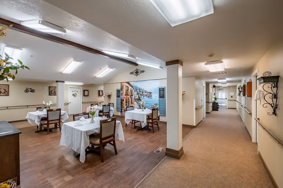 Interior view of a senior living facility dining area with several tables covered in white tablecloths and decorated with vases of flowers. The dining area has wooden flooring and is adjacent to a carpeted hallway with handrails on both sides. The walls are decorated with artwork, including a large mural depicting a scenic coastal village.