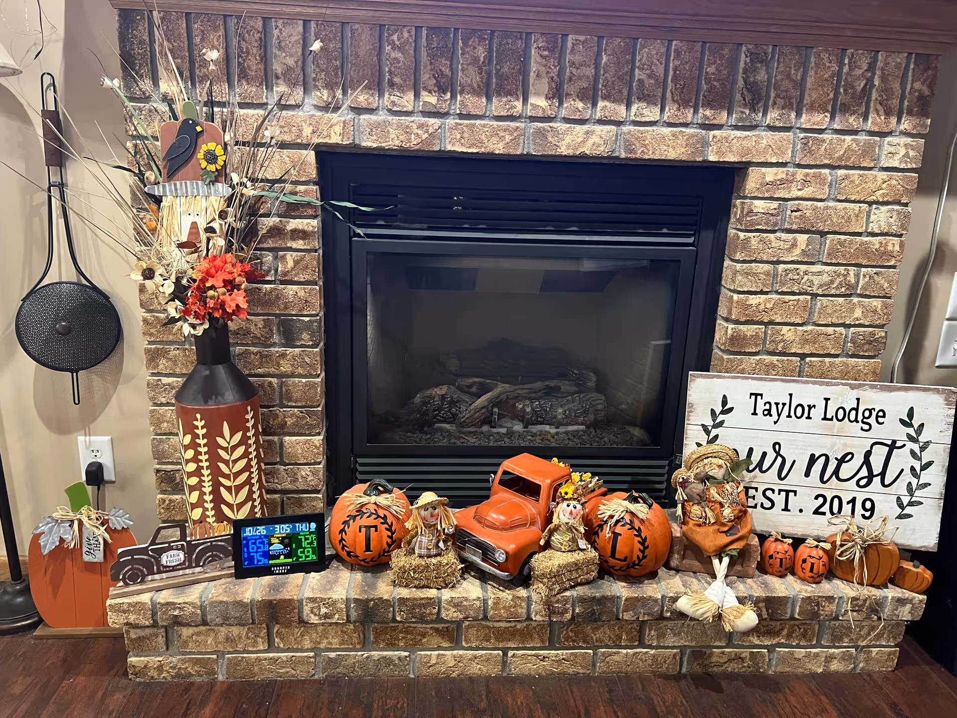 A brick fireplace decorated with autumn-themed items including pumpkins, scarecrows, a small orange toy truck, and a vase with dried flowers. A sign on the right side reads 'Taylor Lodge our nest EST. 2019'. A digital weather station is placed on the hearth among the decorations.