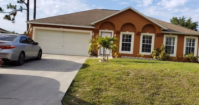 Single-story house with a brown roof and orange exterior walls, featuring three arched windows and a white garage door. A silver car is parked on the driveway, and there is a small palm tree and some shrubs in the front yard with a grassy lawn.