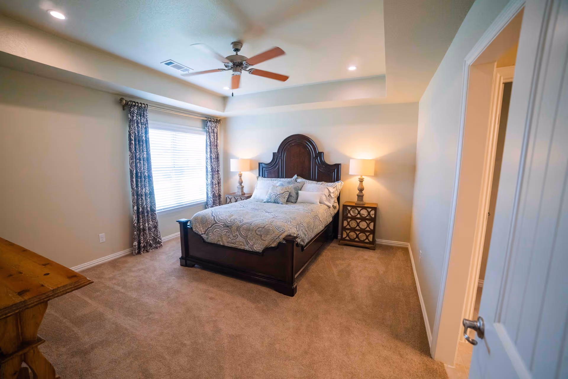 A cozy bedroom with a large dark wooden bed frame and a patterned bedspread. Two bedside tables with matching lamps flank the bed. A window with patterned curtains allows natural light into the room. The ceiling has a fan with wooden blades and recessed lighting. The room has beige carpet and light-colored walls.