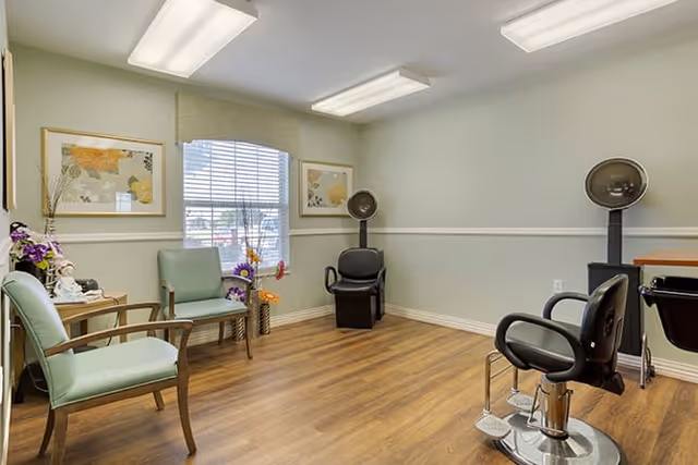 A small salon room with two black salon chairs and hair dryers, two light green chairs, a window with blinds, floral decorations, and framed artwork on the walls. The room has wood flooring and soft green walls with white trim.