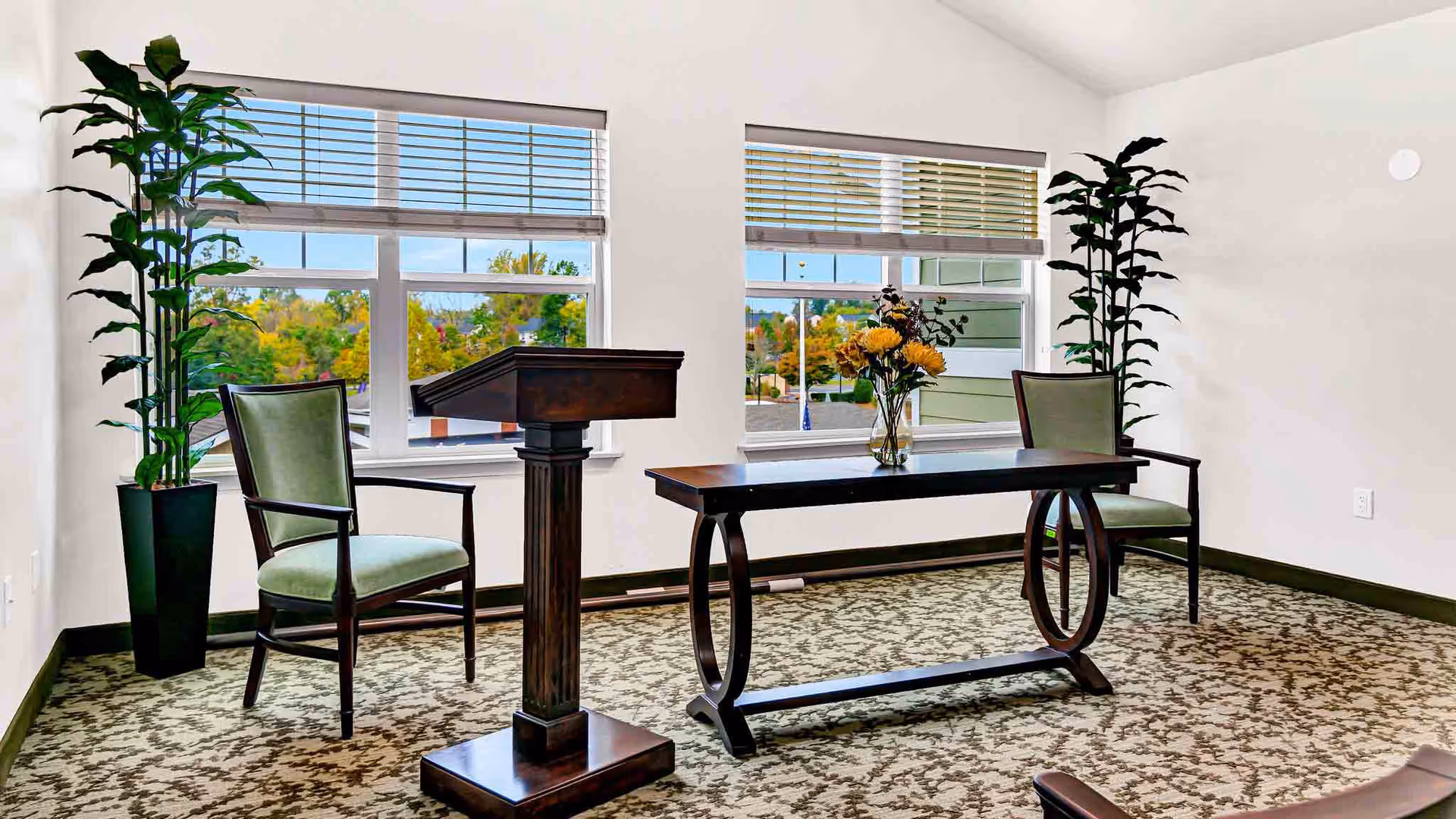 A bright room with two large windows showing an outdoor view with trees. Inside, there are two green cushioned chairs, a dark wooden podium, and a matching table with a vase of flowers. Two tall potted plants are placed near the windows, and the floor is covered with a patterned carpet.